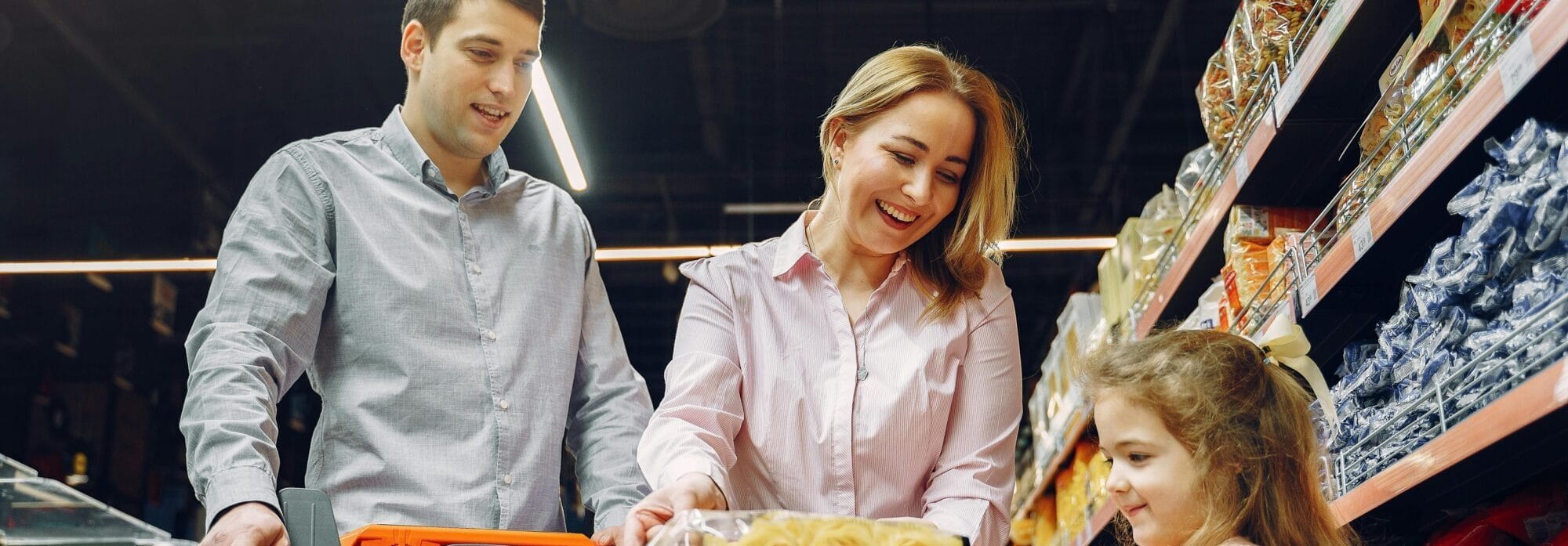 a little girl puts packaged food items in cart as mom and dad push the cart and help her.